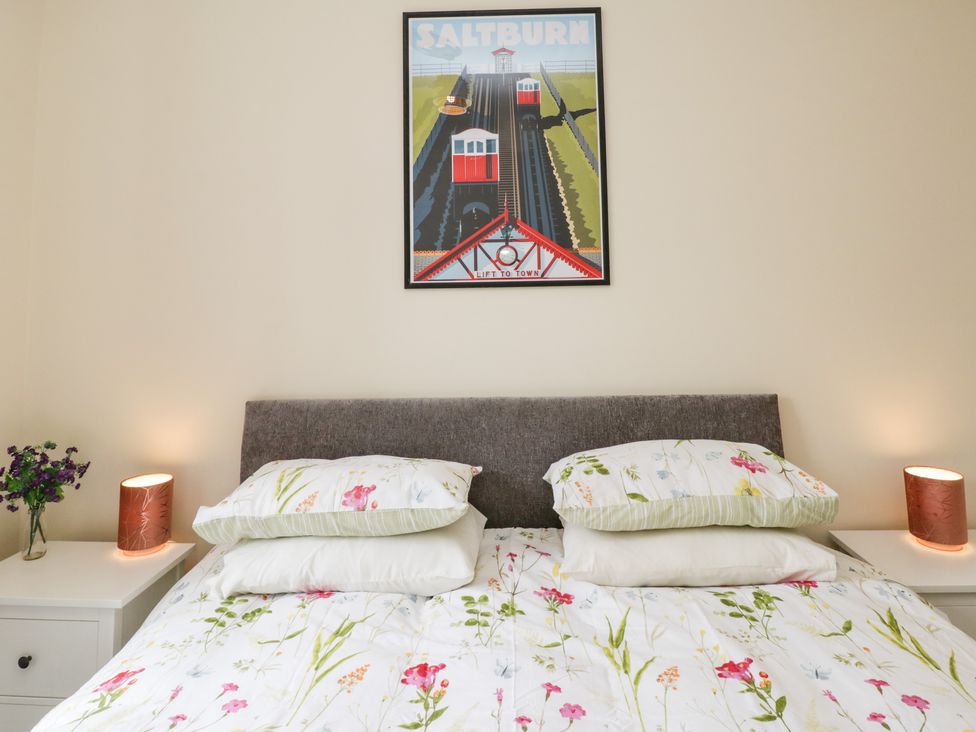 A bedroom with a bed with floral bedding two pillows on each side two bedside tables with lamps and a vase with flowers a framed picture above the bed at Flat 3 in Loftus