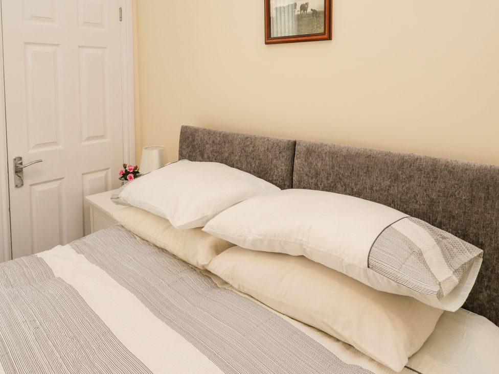 A bedroom with a bed featuring a gray headboard two pillows a bedside table with a lamp and pink flowers a white door and a picture frame on the wall at Flat 3 in Loftus