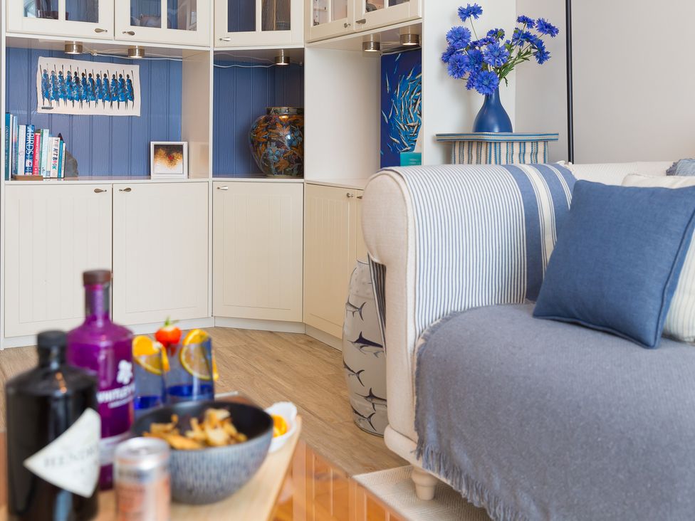 A living room with a sofa covered with blue fabric and cushions a vase with blue flowers on a side table and built in white shelves at Huers Hide Carbis Bay