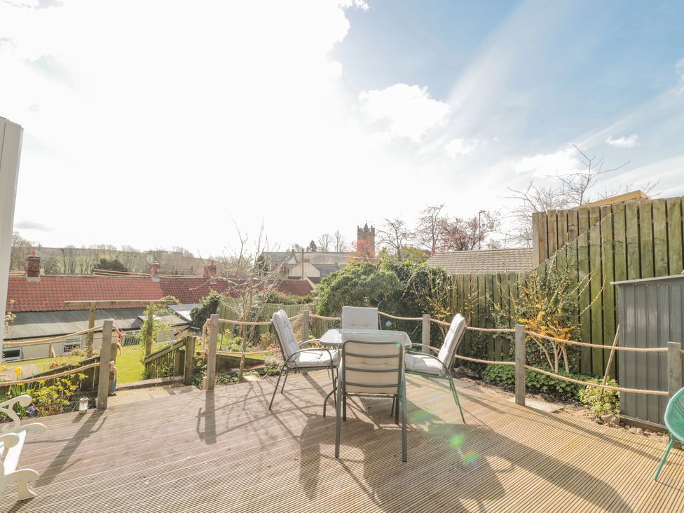 An outdoor wooden deck with four cushioned chairs around a glass table and a wooden fence with plants at Woolley Lodge in Wooler
