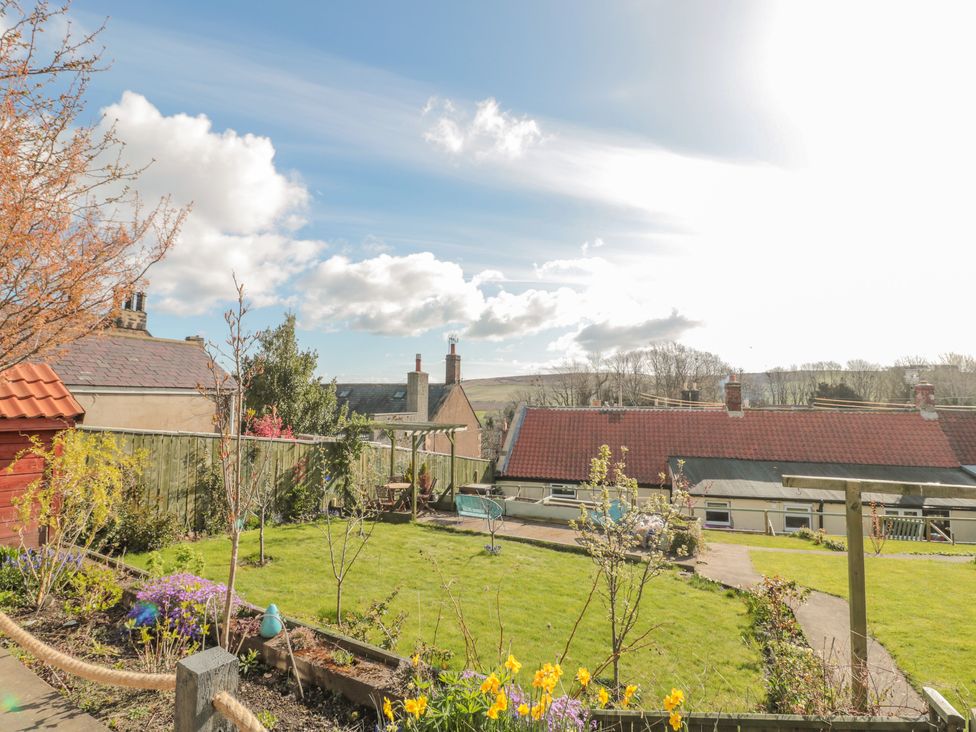 A garden with grass, flowers, and small trees with houses and hills in the background at Woolley Lodge in Wooler