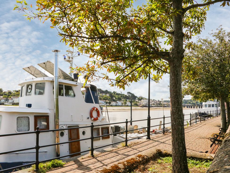 A riverside walkway with trees benches and boats docked by a railing at Courtyard Cottage in Chittlehampton