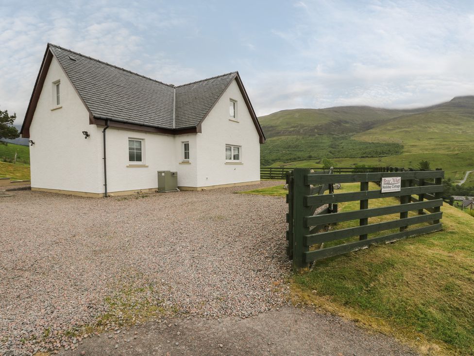A white house with a grey roof on a gravel driveway beside a green wooden gate and hills in the background at Brae Mhor Cottage in Fort William