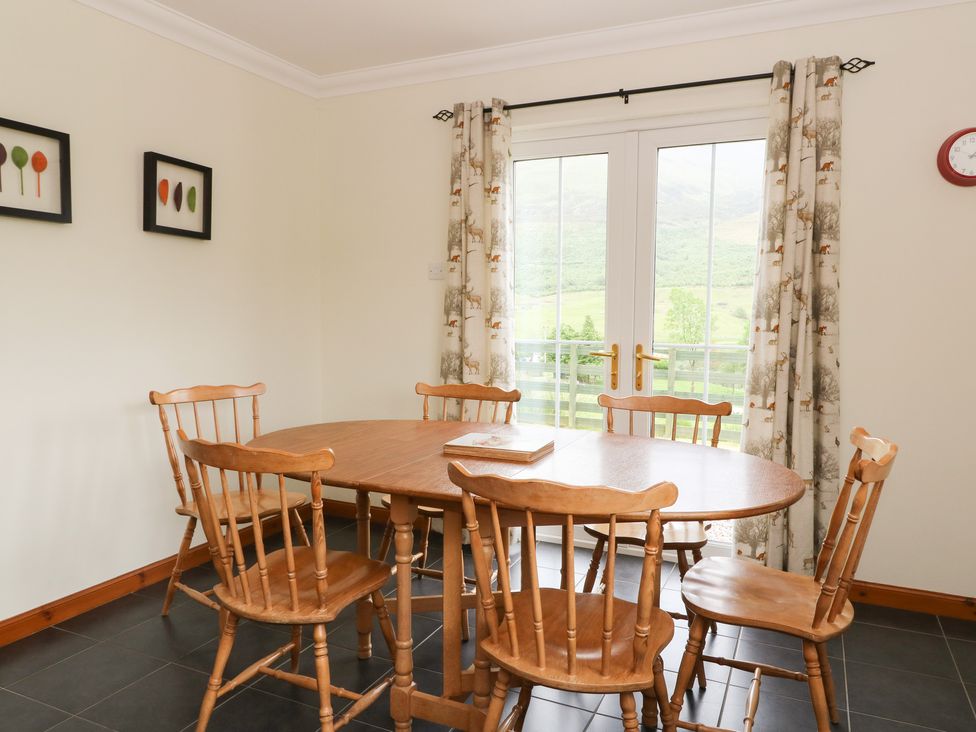 A dining room with wooden table and chairs near glass doors with curtains at Brae Mhor Cottage in Fort William