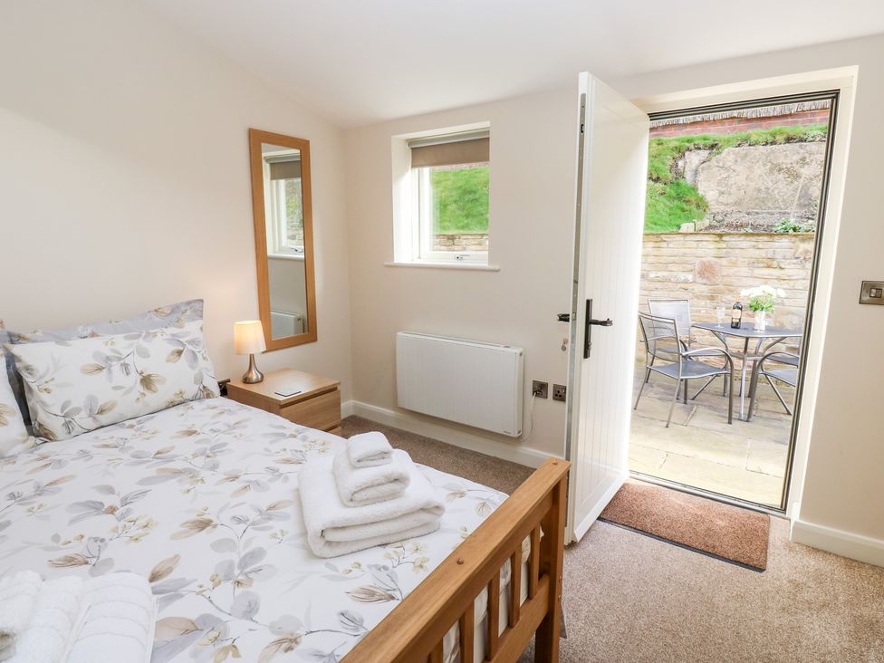 A bedroom with a wooden bed, bedside table with a lamp, a mirror on the wall, an open door leading to a patio with a table and chairs at 2 Barn Cottages in Norwood Green