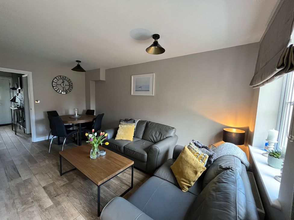A living room with two grey sofas yellow cushions a wooden coffee table with flowers a dining table with black chairs and a wall clock at Gone Crabbing in The Bay Filey