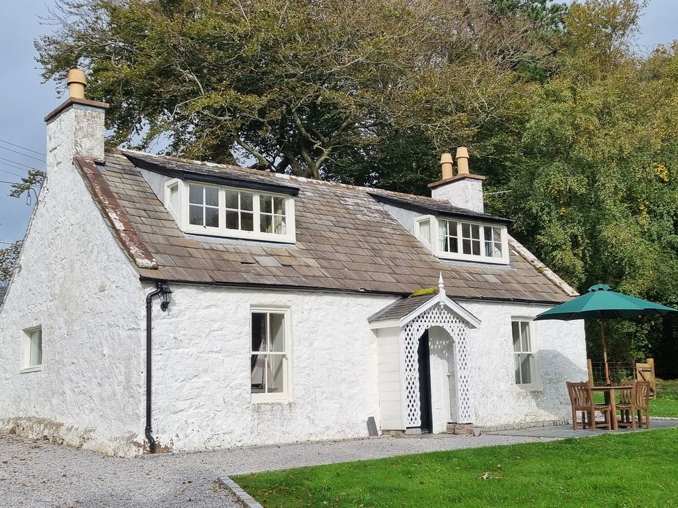 A white stone cottage with two chimneys and dormer windows surrounded by trees with outdoor table and umbrella at Saltflats Cottage Rockcliffe near Dalbeattie