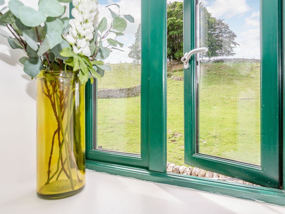 A yellow vase with flowers on a windowsill with a green window frame and a view of a grassy field and trees at Pogles Wood Cottage in Grassington