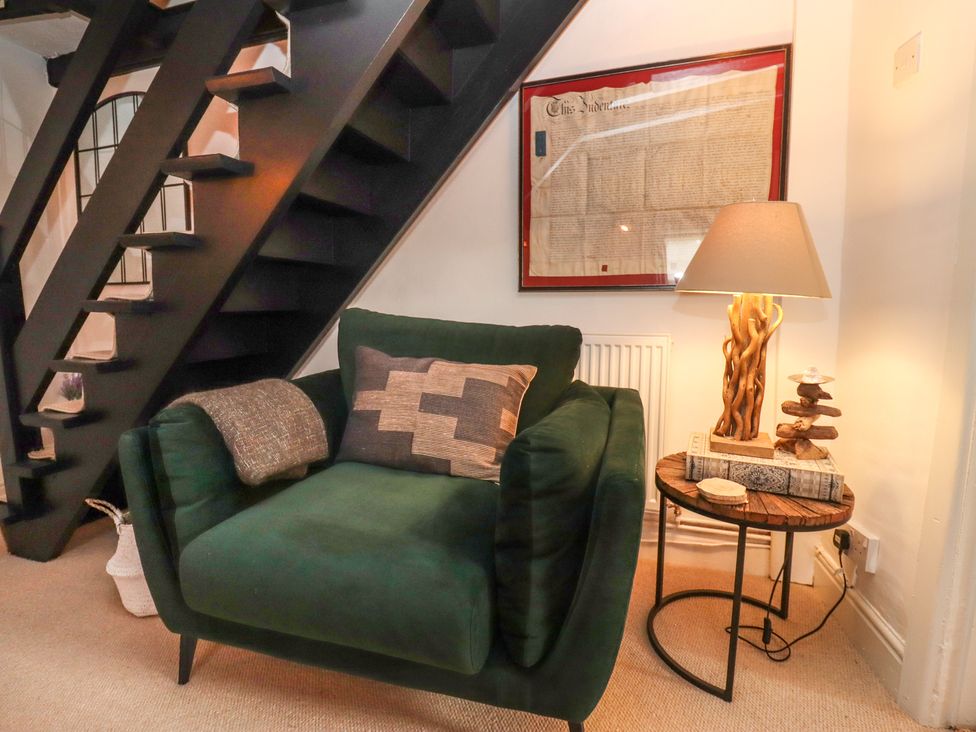 A green armchair with cushions next to a wooden side table with a lamp and stacked books under a staircase at Hobbes Cottage in Malmesbury