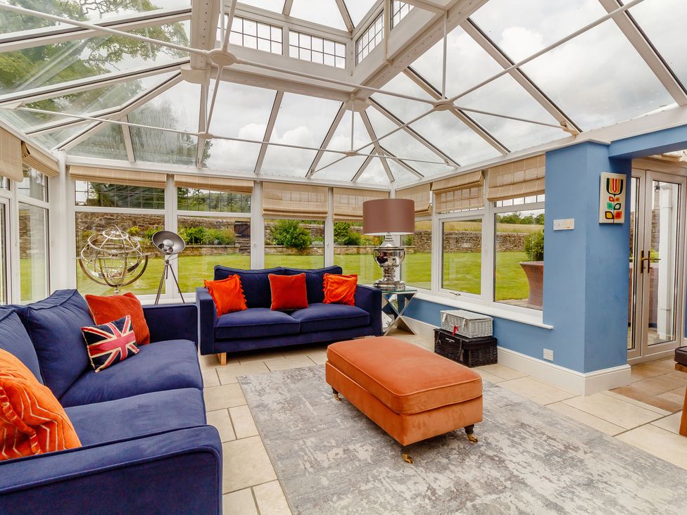 A conservatory with blue sofas orange cushions an orange ottoman a floor lamp and large windows at Fence House in Fence