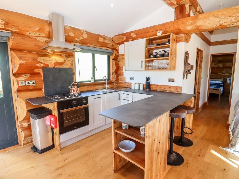 A kitchen with wooden walls white cabinets a stove and oven a sink a countertop with stools and an open door showing a bedroom at Cedar Cabin Near Clifton Upon Teme
