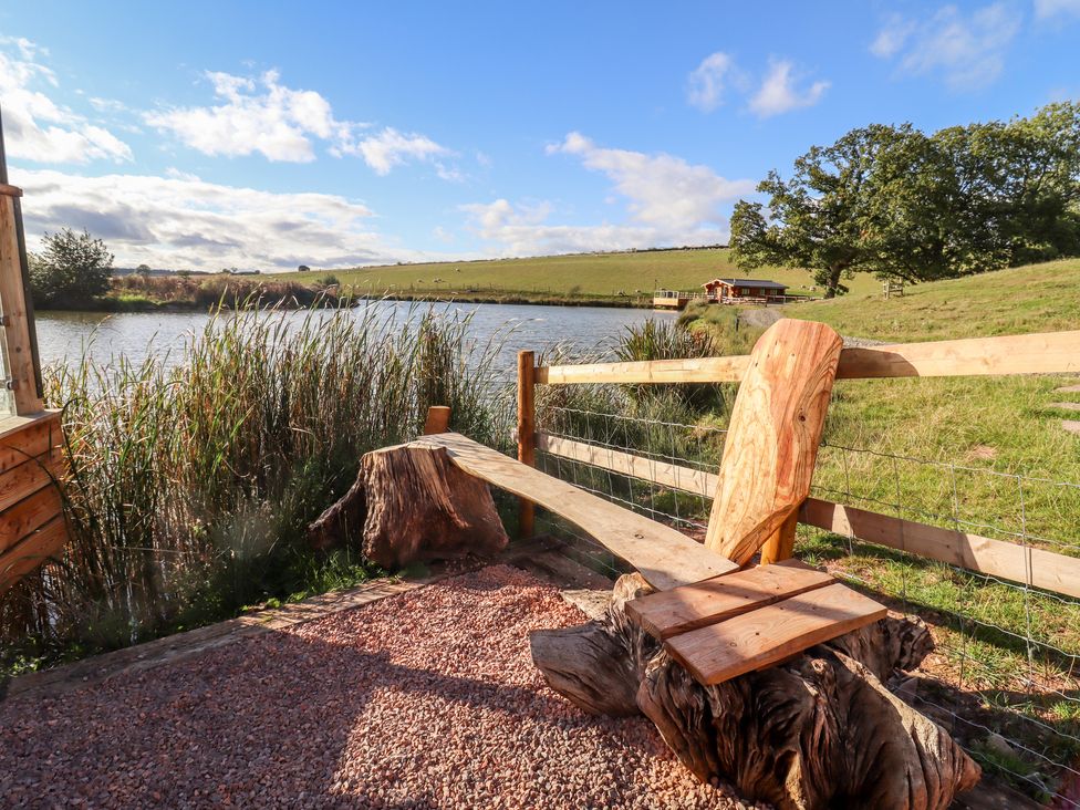 A wooden bench made from tree logs near a lake surrounded by reeds and grass at Cedar Cabin Near Clifton Upon Teme
