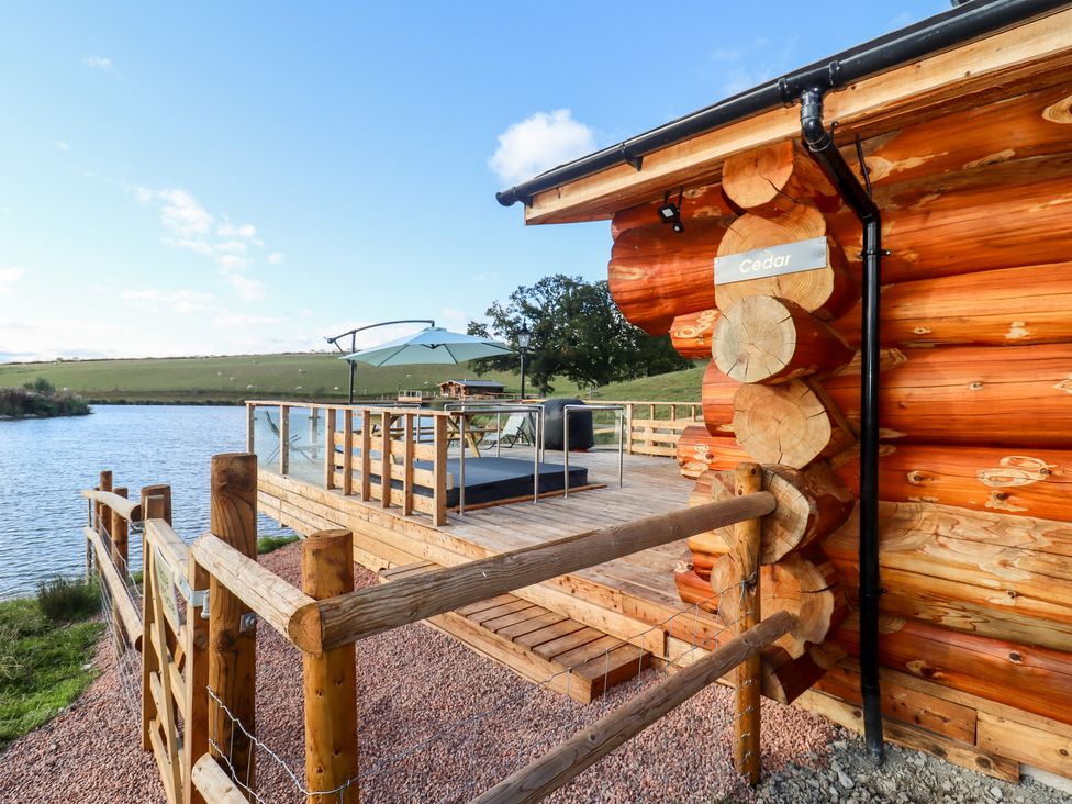 A wooden deck with railing and an umbrella near a lake next to a log cabin at Cedar Cabin Near Clifton Upon Teme