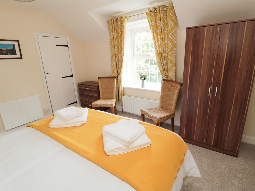 A bedroom with a bed covered by a yellow blanket and towels on top two wicker chairs near a window with yellow patterned curtains a wooden wardrobe and a chest of drawers at 1 Roddam Rigg Cottage Roddam near Wooler