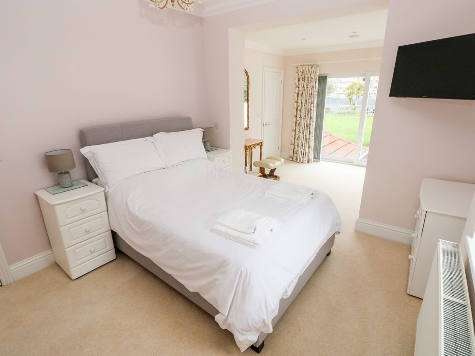 A bedroom with a bed and white bedding two bedside tables lamps a wall mounted tv and glass doors with curtains at The Headlands - Gower in The Mumbles