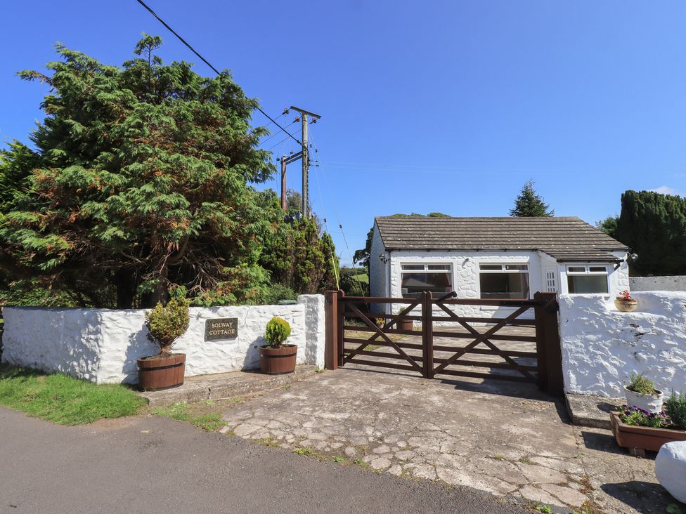 A white stone cottage with a gated driveway and large trees behind a white wall at Solway Cottage Powfoot near Annan