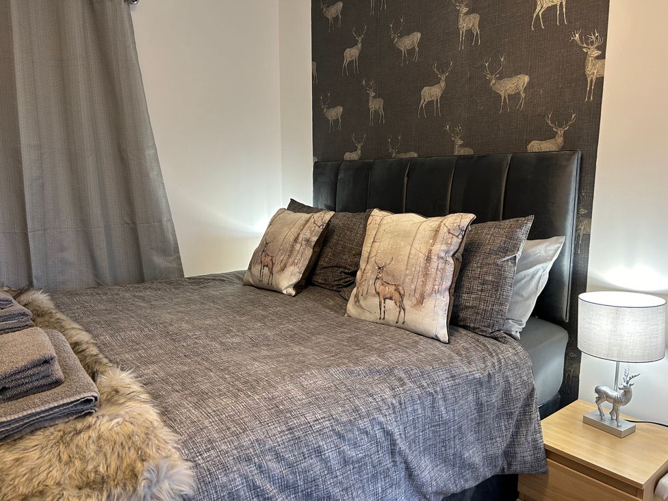 A bedroom with a bed covered with a gray bedspread and cushions with deer prints a bedside table with a lamp and folded towels on a fur rug at Solway Cottage in Powfoot near Annan