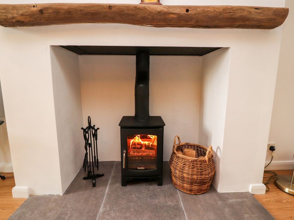 A wood-burning stove with fire inside a recessed fireplace with a basket of firewood and fireplace tools at Top House in Settle
