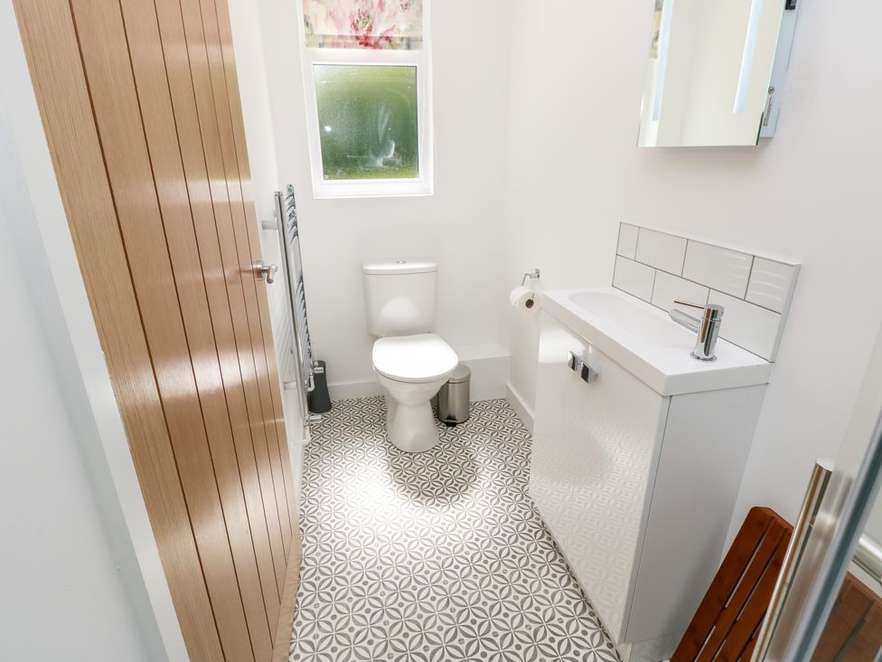 A bathroom with a toilet, a sink with cabinet, a mirror, a wooden door, a window with a floral blind, and patterned floor tiles at Cava in Seaview