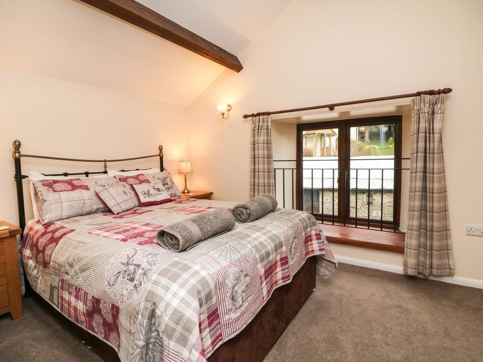 A bedroom with a double bed with patterned bedding beside a window with curtains and wooden furniture at Red Deer Cottage in Wheddon Cross