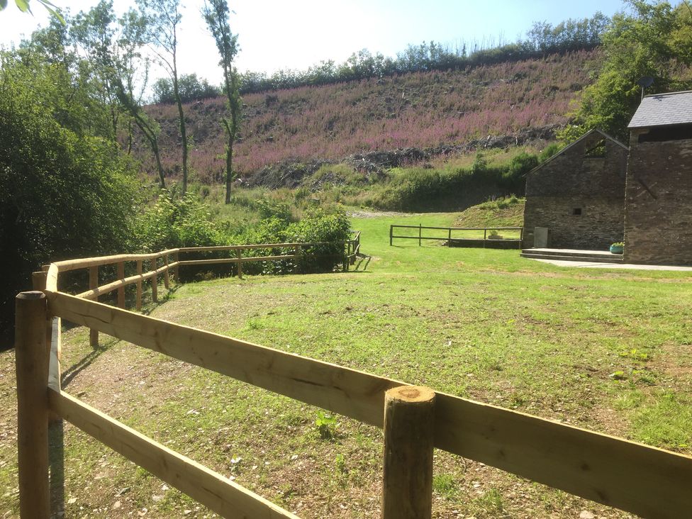 An outdoor area with grass a wooden fence trees and a stone building at Red Deer Cottage in Wheddon Cross