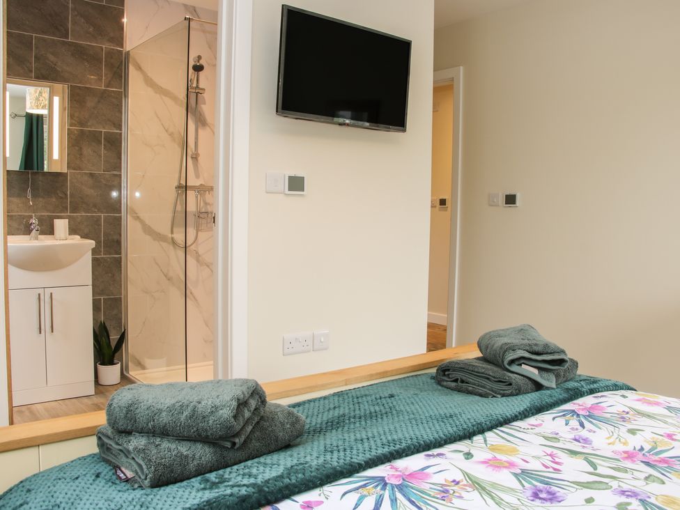 A bedroom with a bed and folded towels facing a wall-mounted television and an open doorway showing an en suite bathroom with a shower and sink at Holly Lodge in Forden near Montgomery