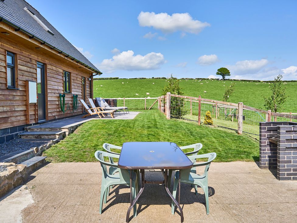 An outdoor patio with a table and chairs next to a wooden house and a grassy fenced yard in a rural area at Holly Lodge in Forden near Montgomery