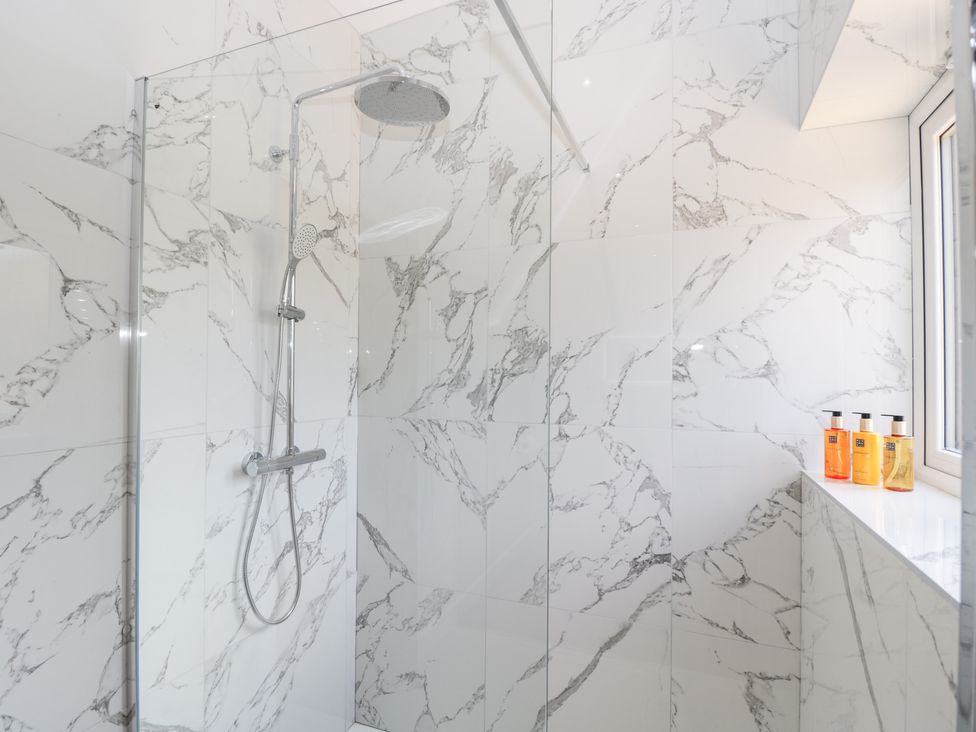 A shower area with a rainfall showerhead and a handheld shower on marble tiled walls with bottles on the windowsill at Stag Cottage in Lytchett Matravers