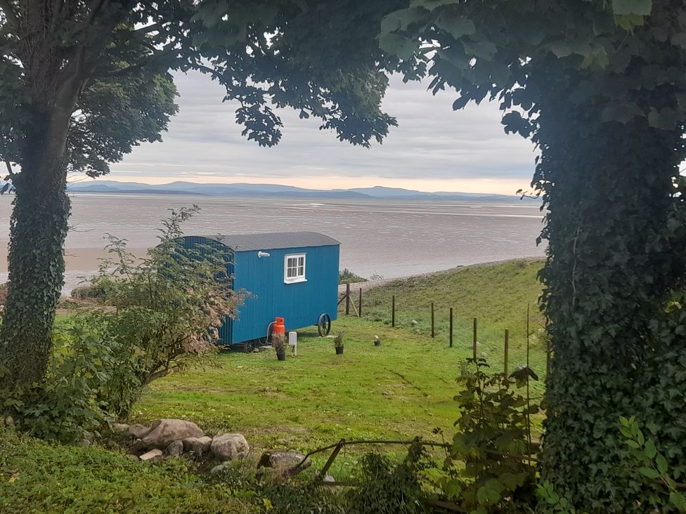 A blue shepherds hut on grass near a body of water with trees in the foreground at Seashore Shepherds Hut @ Moat Farm in Aldingham near Ulverston