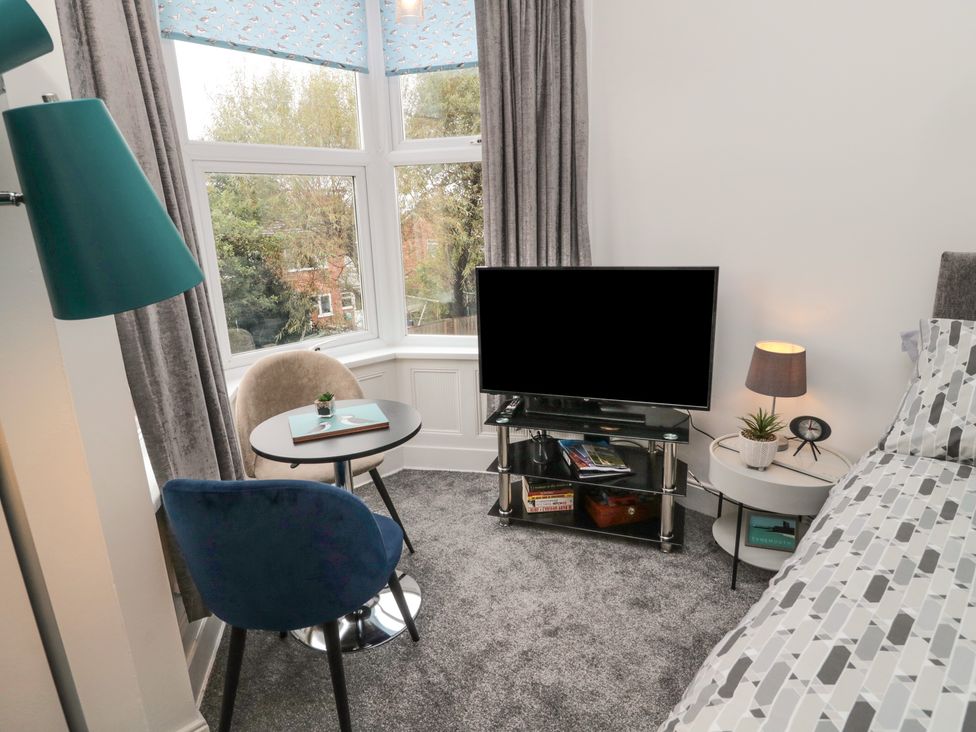 A bedroom with a bed next to a round side table with a lamp and clock a TV on a stand and a small round table with two chairs near a window at Studio 51 @ Tynemouth in Tynemouth