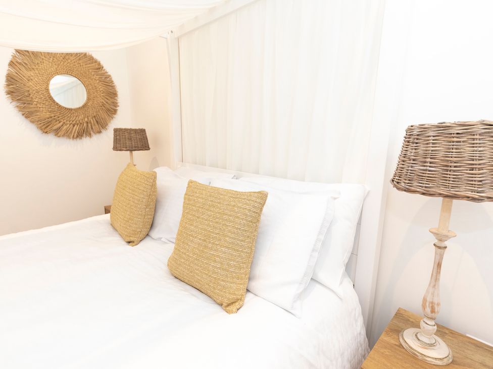 A bedroom with a white bed with two yellow pillows a wicker lamp on a wooden nightstand and a round wicker mirror on the wall at Bishops Retreat in Ventnor