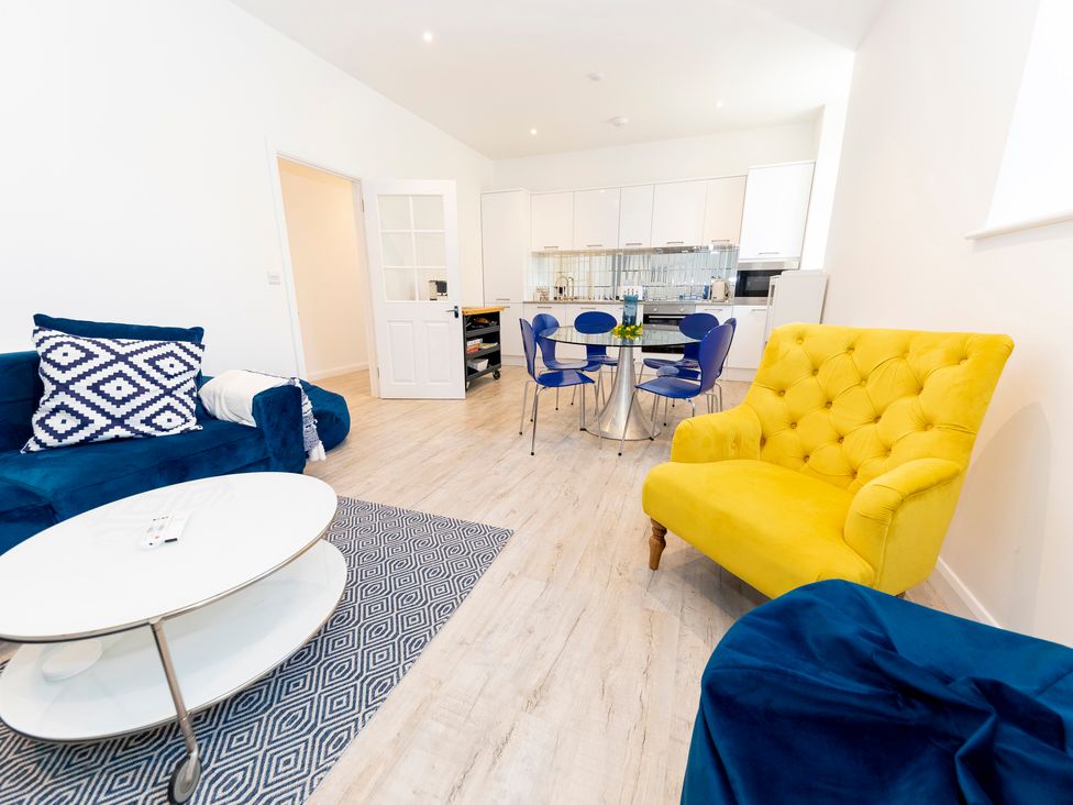 A living area with blue sofa yellow armchair glass dining table with blue chairs and open kitchen in the background at Bishops Retreat in Ventnor