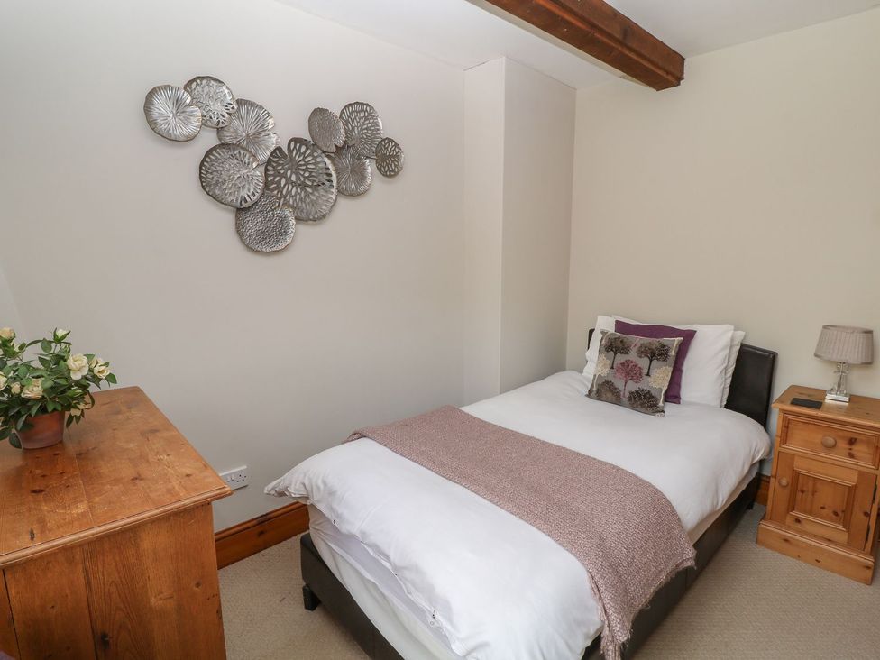 A bedroom with a single bed decorative pillows a wooden nightstand with a lamp a wooden dresser with a plant and metal wall art at Hazel Barn North Molton