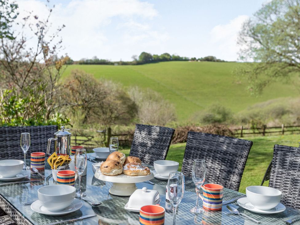 An outdoor dining table set with bowls mugs a teapot and pastries with chairs and a green field in the background at Hazel Barn in North Molton