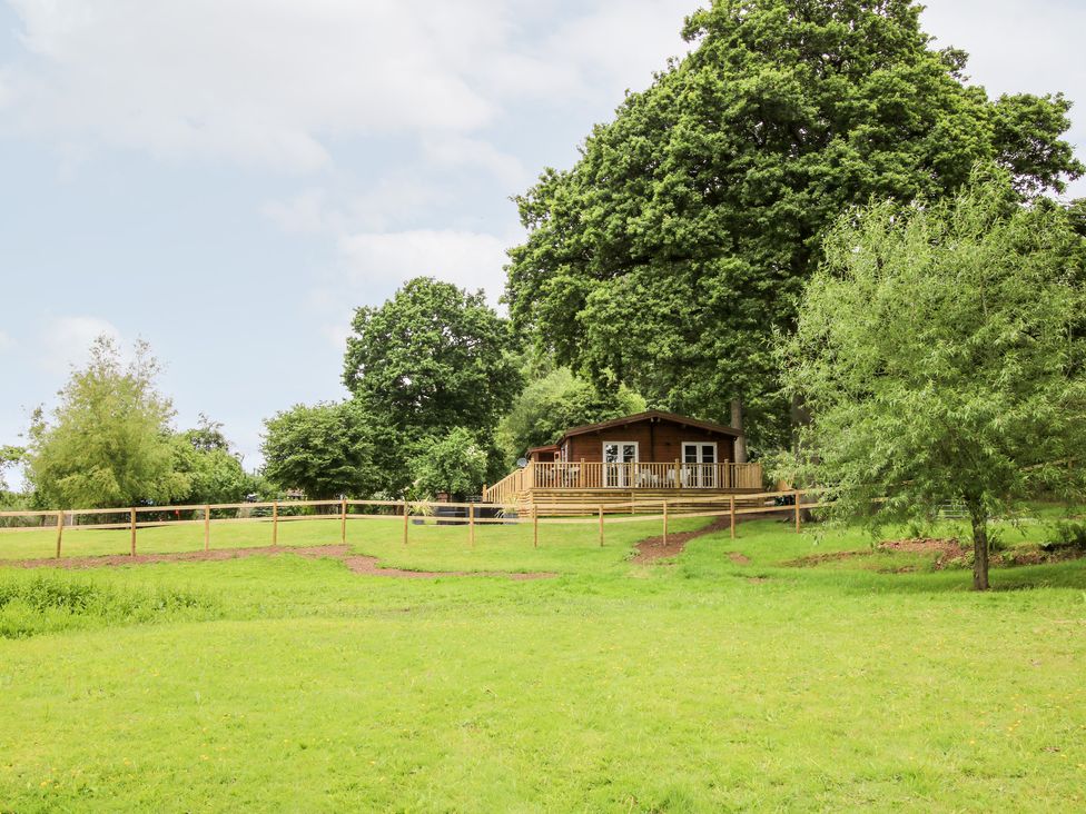 A wooden cabin with a deck surrounded by trees and a fenced grassy area at The Log Cabin in Newnham Bridge near Tenbury Wells