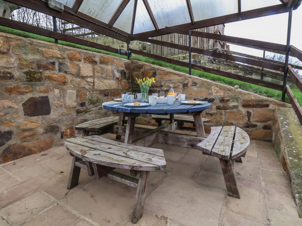 A wooden picnic table set with cups plates and a vase of flowers under a transparent roof with stone walls around at The Barn in Staithes