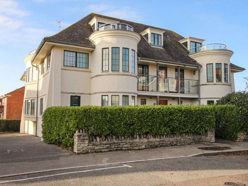 A white two-story house with curved bay windows and a glass balcony behind a hedge at Broadleys in Swanage