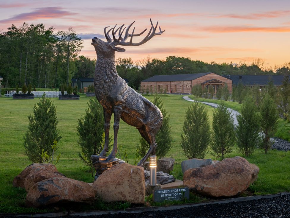 A metal stag statue on a grassy area with rocks and small trees near wooden buildings at Lodge 7 - The Earn in Errol