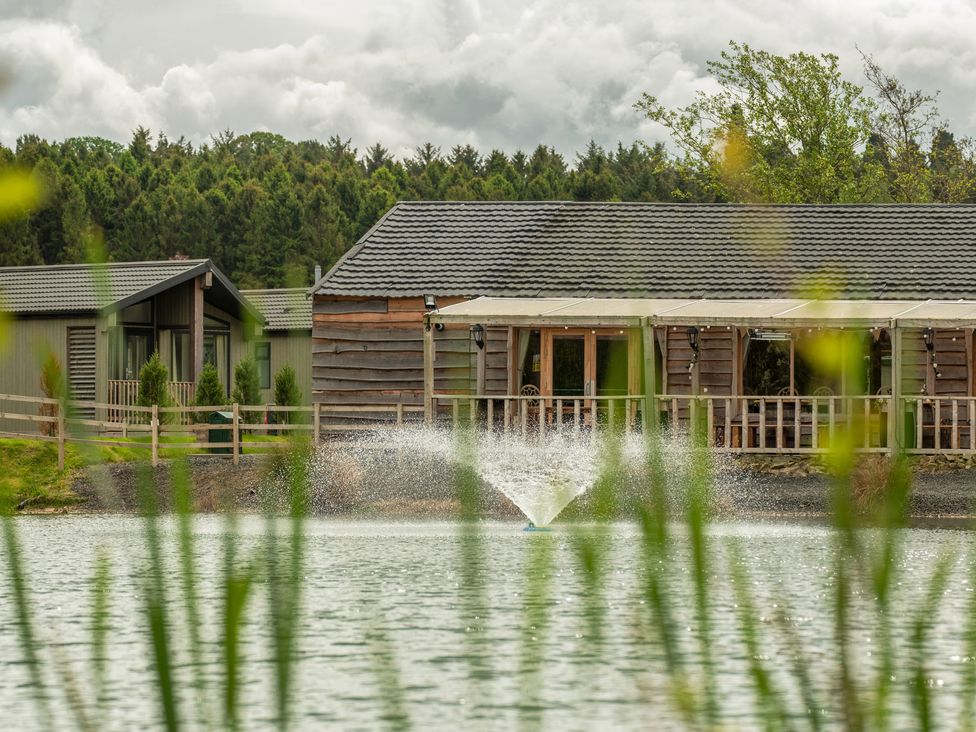 A pond with a water fountain in front of wooden cabins with a railing and trees in the background at Lodge 7 - The Earn in Errol