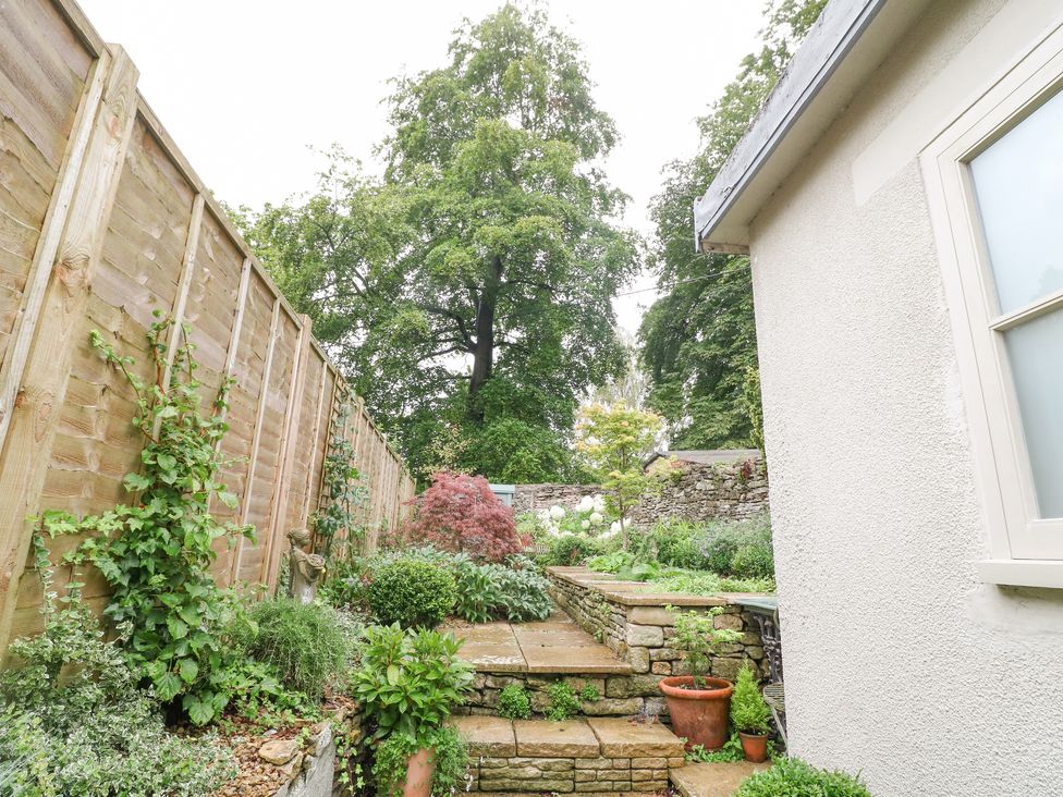 A garden with stone steps leading to plants and trees beside a wooden fence and a house wall at The Snug No 35 West End in Minchinhampton