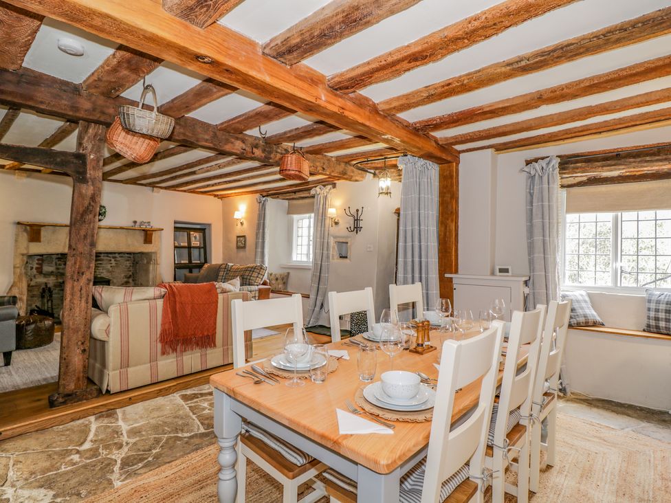 A dining area with a wooden table set with dishes and chairs next to a living room with sofas and a fireplace at Fox Den Cottage in Castle Combe