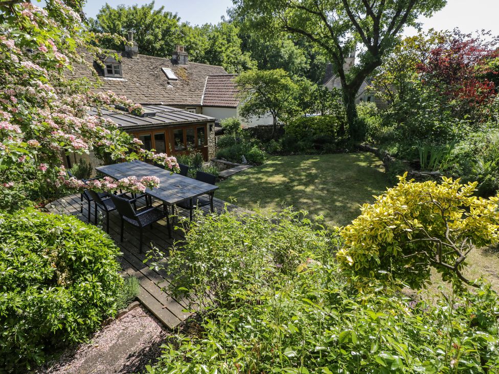 An outdoor wooden dining table with chairs on a wooden deck surrounded by trees and bushes at Fox Den Cottage in Castle Combe
