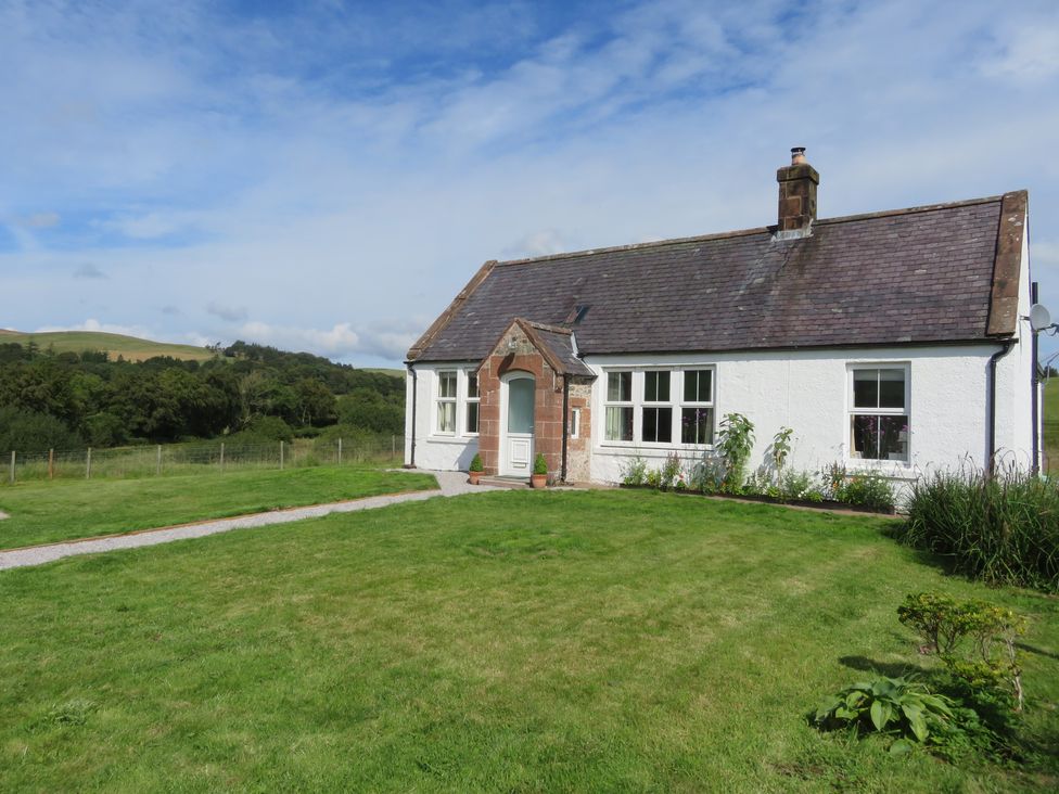 A white cottage with a stone chimney and front door on a grassy lawn with plants and flowers at Marr Cottage in Thornhill