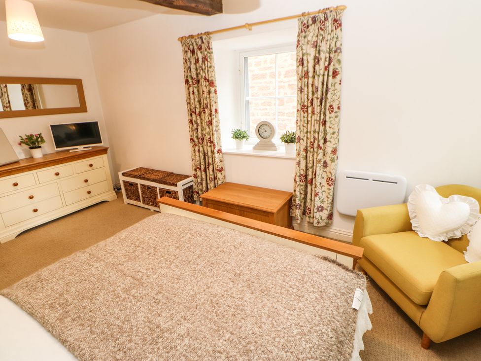 A bedroom with a bed, yellow armchair, dresser with TV, storage baskets, and window with floral curtains at Pine Cottage in Appleby-In-Westmorland