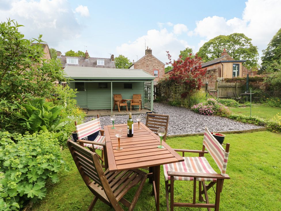 A garden with a wooden table and chairs on grass near a green shed with houses and plants in the background at Pine Cottage in Appleby-In-Westmorland