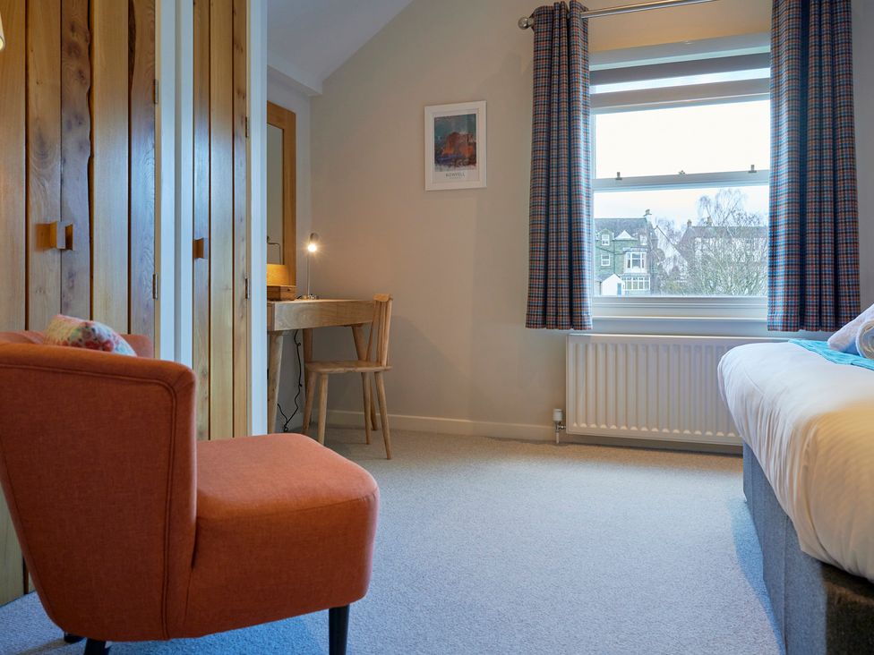 A bedroom with a red chair wooden wardrobe desk and window with curtains at Howe Keld 5 First Floor Keswick