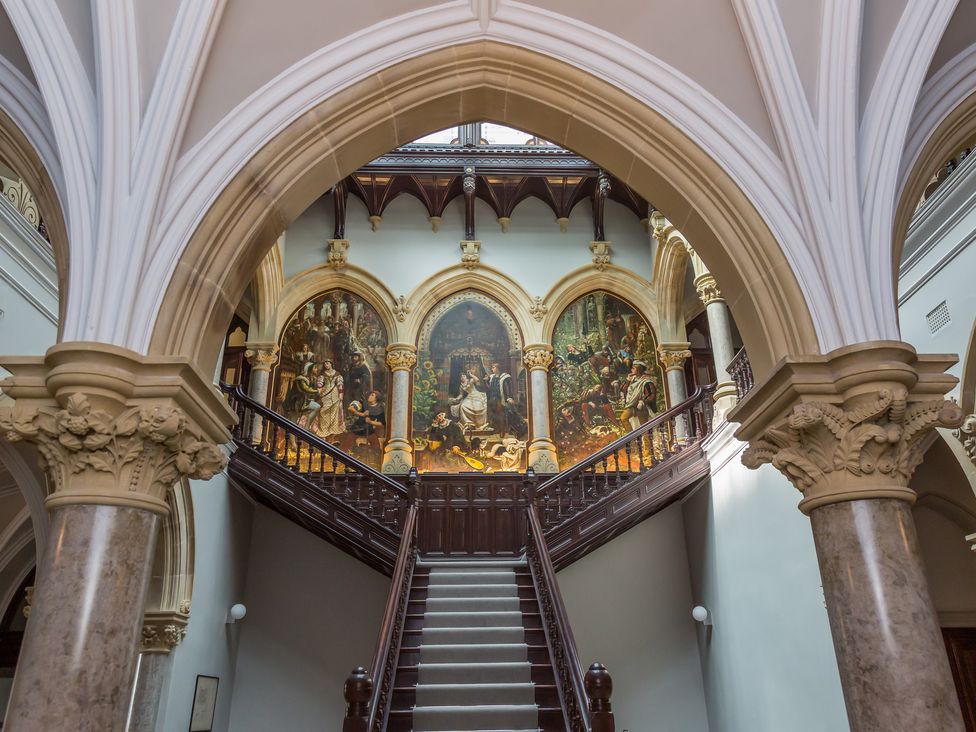 An ornate staircase with three large paintings above and detailed arches around at The Mabel Suite Stone Cross Mansion in Ulverston