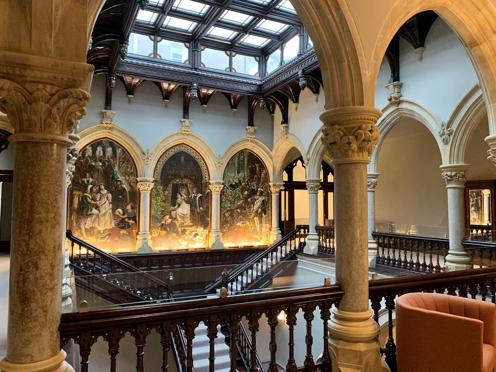 An interior view of a historic building with arched columns a skylight wooden railings and large paintings on the wall at The Mabel Suite Stone Cross Mansion in Ulverston