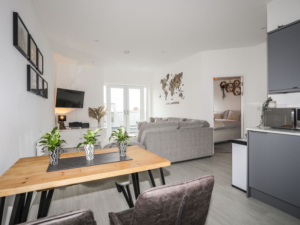 A living room with a wooden table with plants chairs a gray sofa a wall-mounted tv and a kitchen area at Brig-Y-Don 4 in Rhosneigr