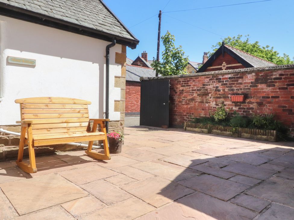 An outdoor patio area with a wooden bench and flower pot against a white wall and a brick wall with plants at The Mews Cottage in Lytham St Annes